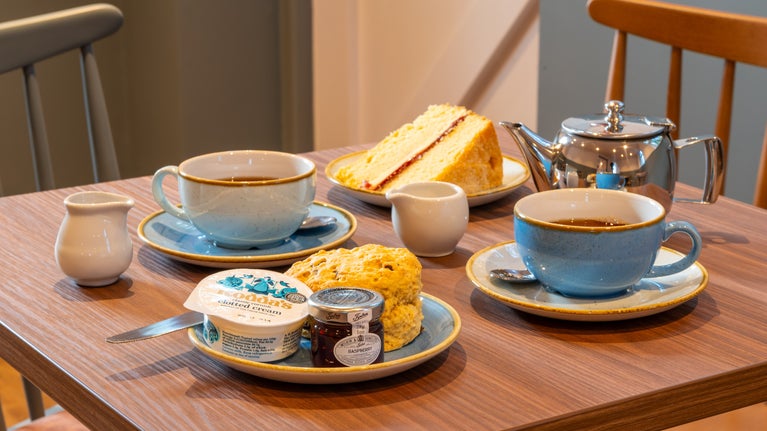A table arranged with 2 cups of tea, tea pot, milk jug, slice of Victoria sandwich cake, a plate with a scone and pot of cream and jam